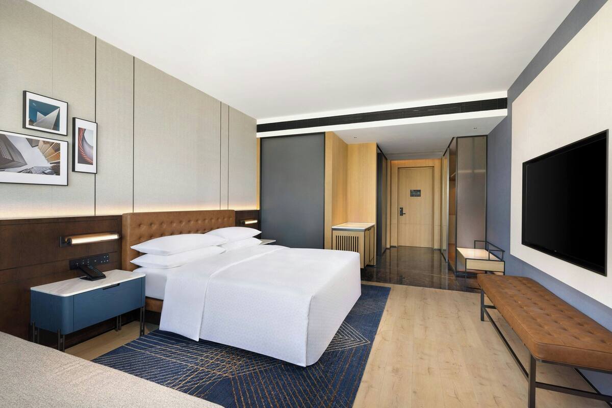 A modern hotel room with a large bed, bedside tables, a flat-screen TV, and a bathroom visible in the background.