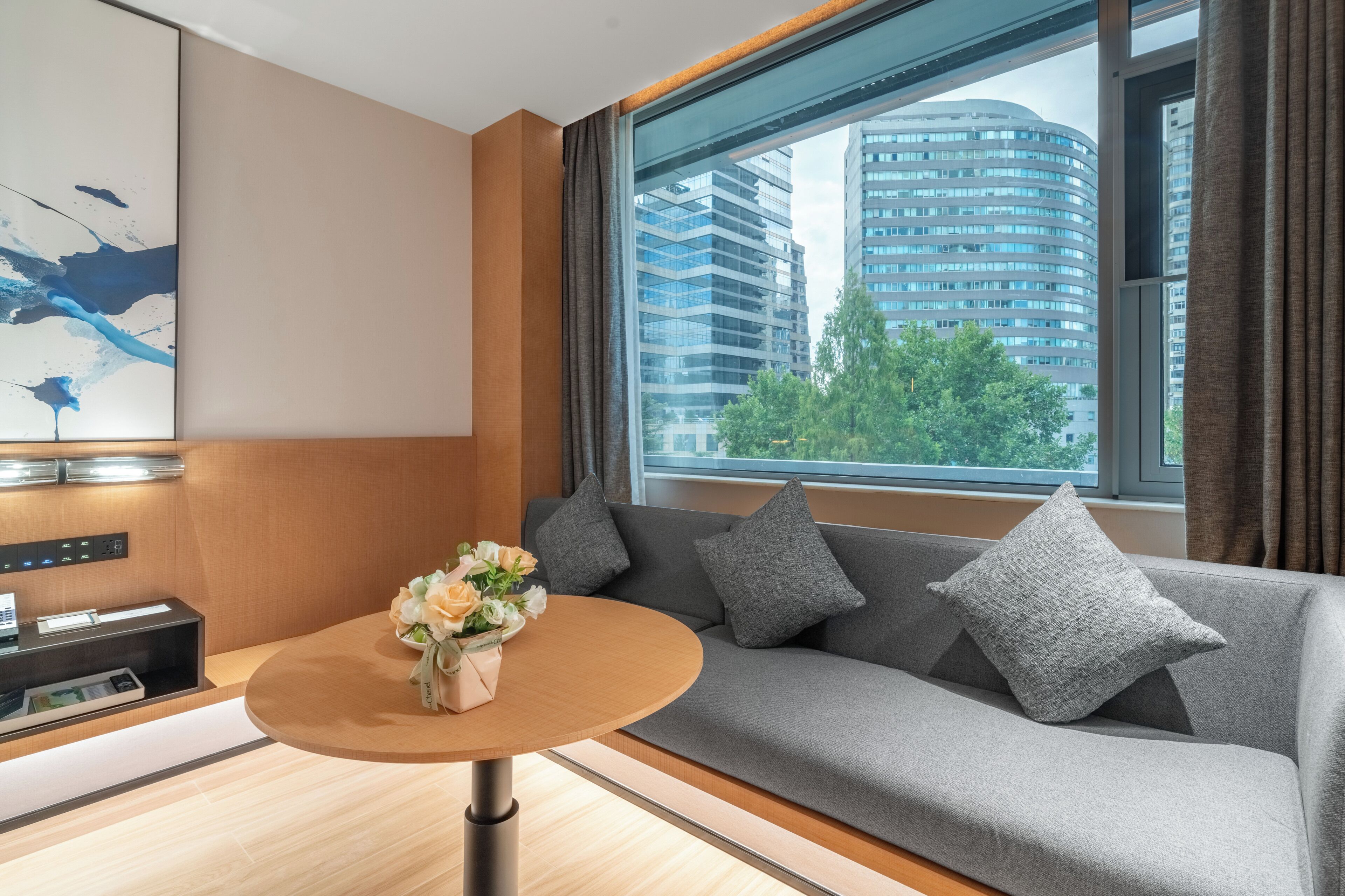 A modern hotel room with a sofa, a round table with a vase of flowers, a control panel, and a large window with a city view.
