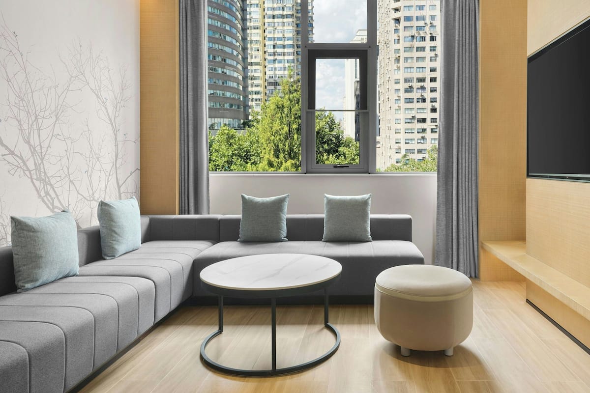 Ein modernes Wohnzimmer mit einem grauen Ecksofa, einem runden Couchtisch und einem kleinen Hocker. Ein großes Fenster bietet Blick auf hohe Gebäude und Bäume.