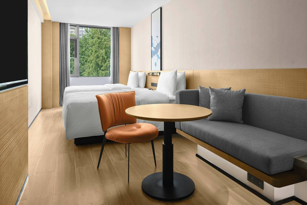 Ein modernes Hotelzimmer mit Bett, Sofa, rundem Tisch und Blick auf Grün hinter dem Fenster.