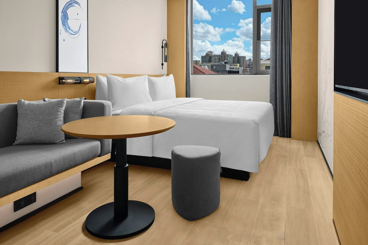 Ein modernes Hotelzimmer mit einer Couch, einem Einzelbett, einem kleinen runden Tisch und Blick auf die Stadt durch ein großes Fenster.