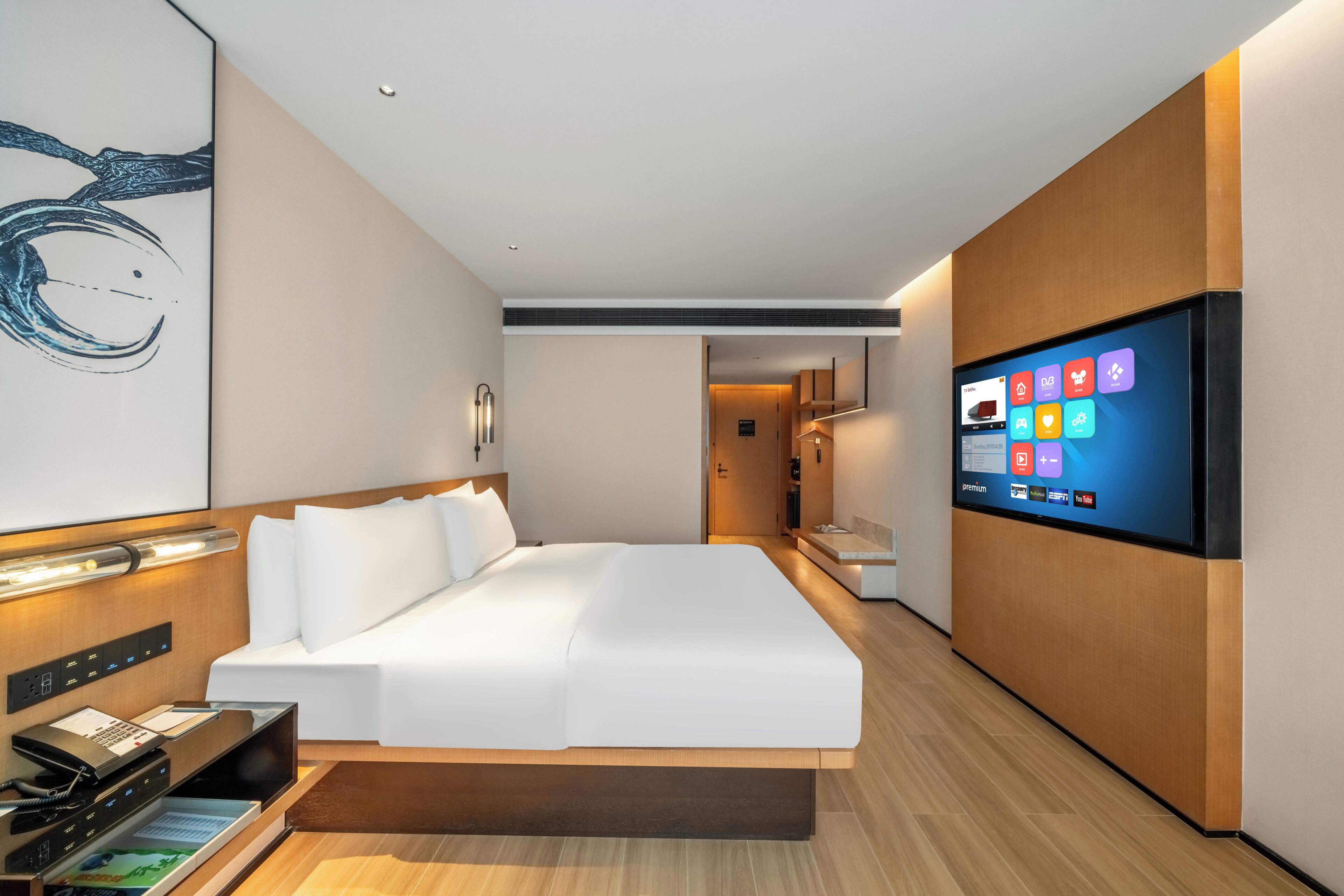 A modern hotel room with a bed, a flat-screen TV, a desk with a phone, and a wall-mounted artwork.