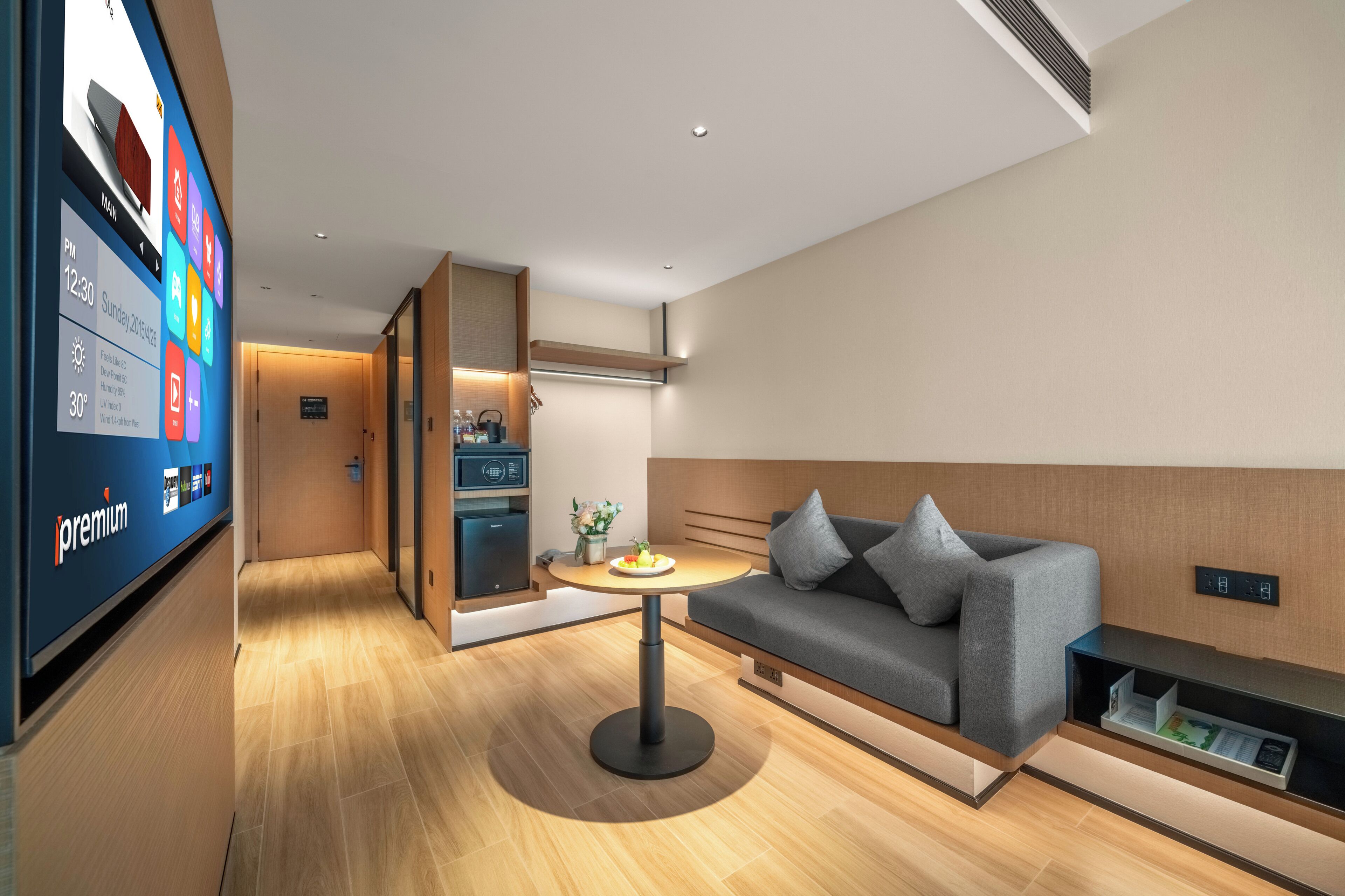 A modern hotel room with a flat-screen TV displaying room information, a sofa, a small round table, and a minibar.