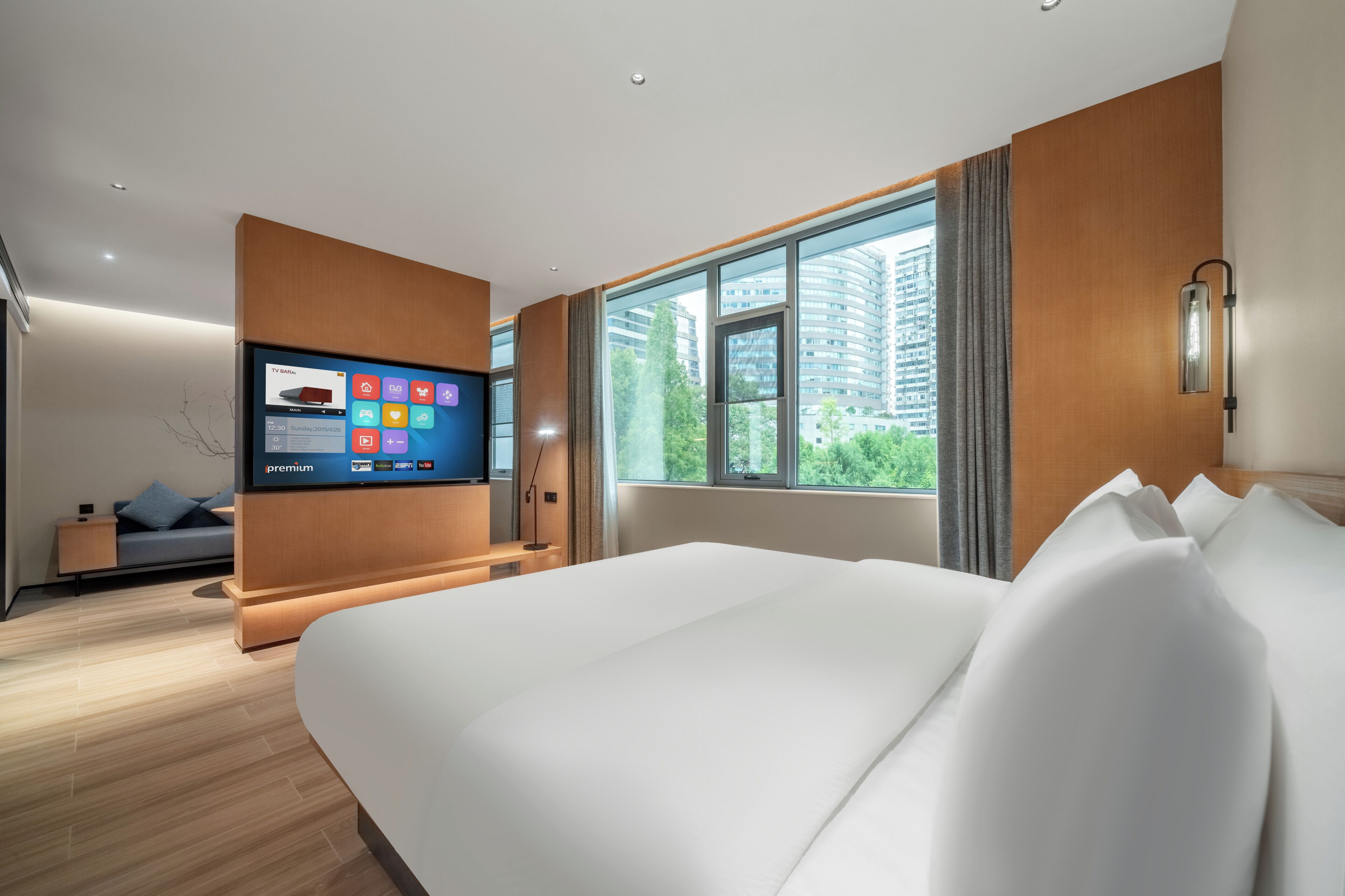 A modern hotel room with a large bed, a TV mounted on the wall, a window with a view of buildings, and a small seating area with a sofa and a side table.