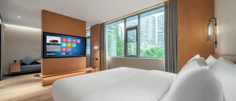 A modern hotel room with a large bed, a TV mounted on the wall, a window with a view of buildings, and a small seating area with a sofa and a side table.