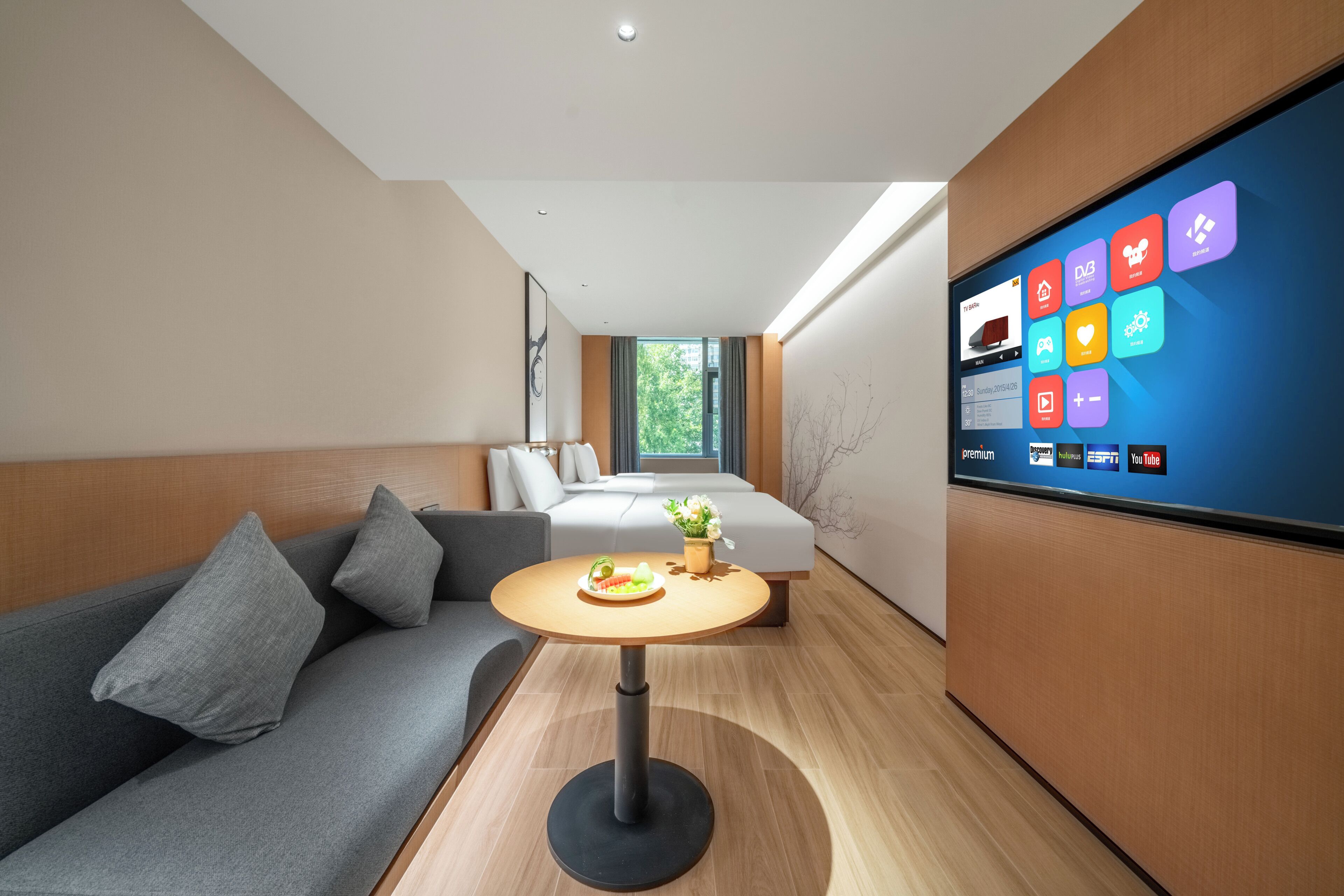 A modern hotel room with a sofa, a small round table with a plate of food, a bed with a view of greenery, and a wall-mounted TV displaying various apps.