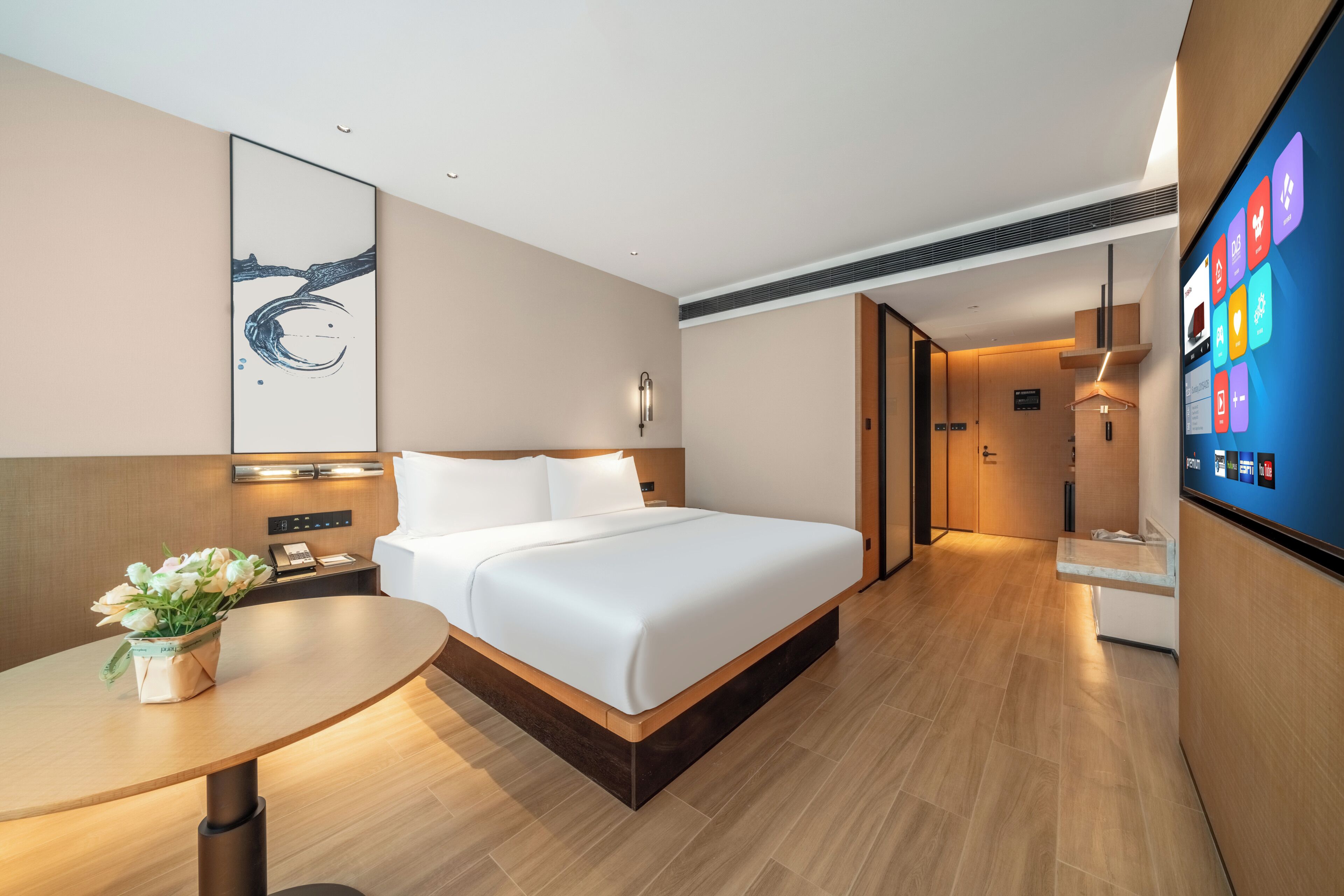 A modern hotel room with a large bed, bedside tables, a desk, and a digital screen displaying room information.