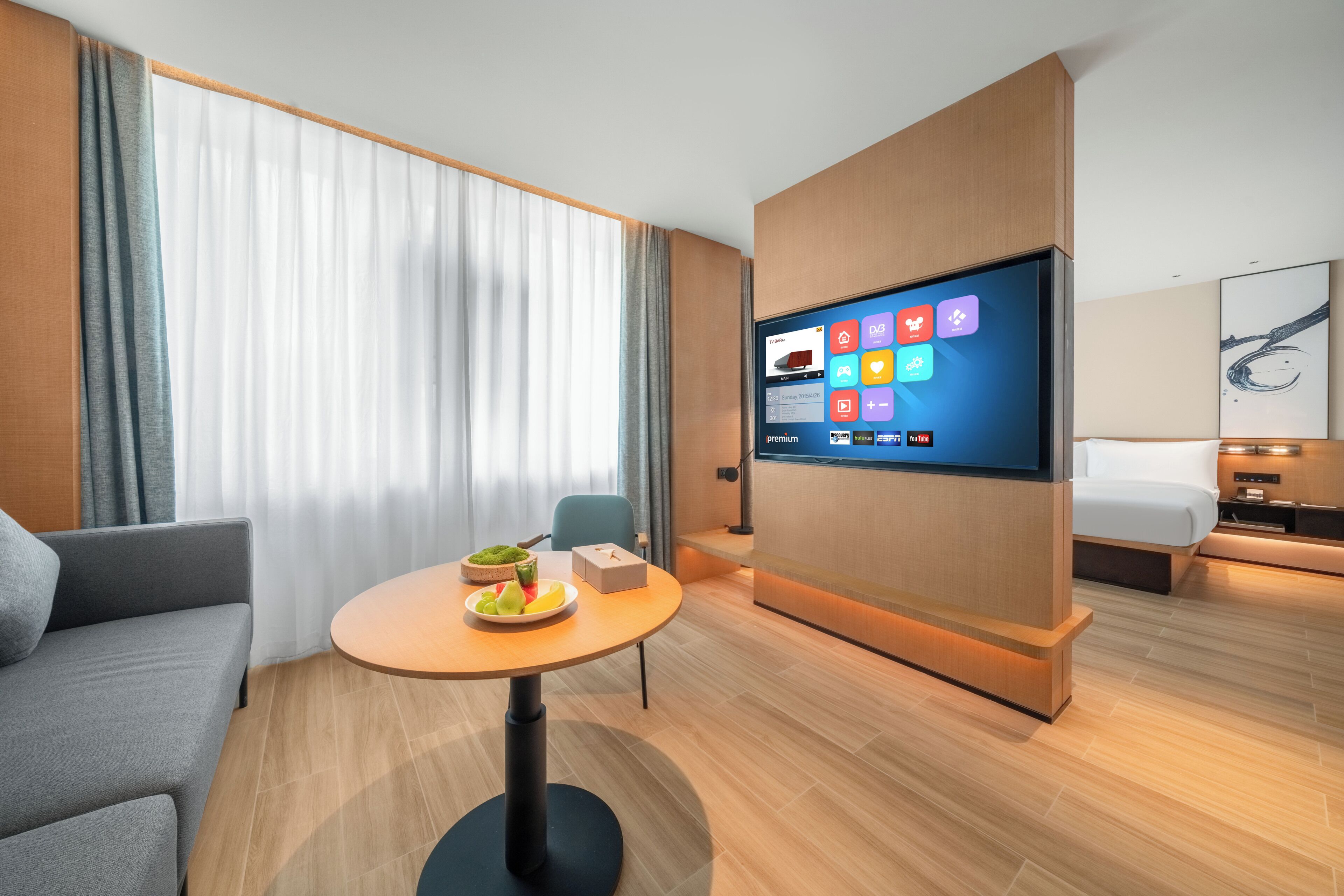 A modern hotel room with a flat-screen TV, a round table with fruit, a sofa, and a bed.