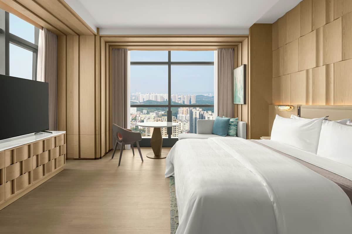 A modern hotel room with a large bed, a TV, a desk, and a view of the cityscape.