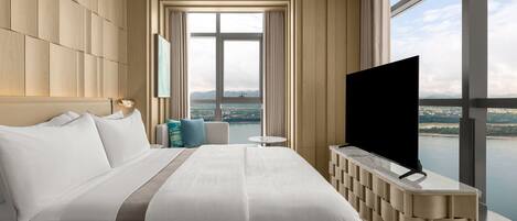 Une chambre d’hôtel avec un grand lit, un téléviseur posé sur une console en bois, une vue sur une étendue d’eau et un panorama de la ville au loin.