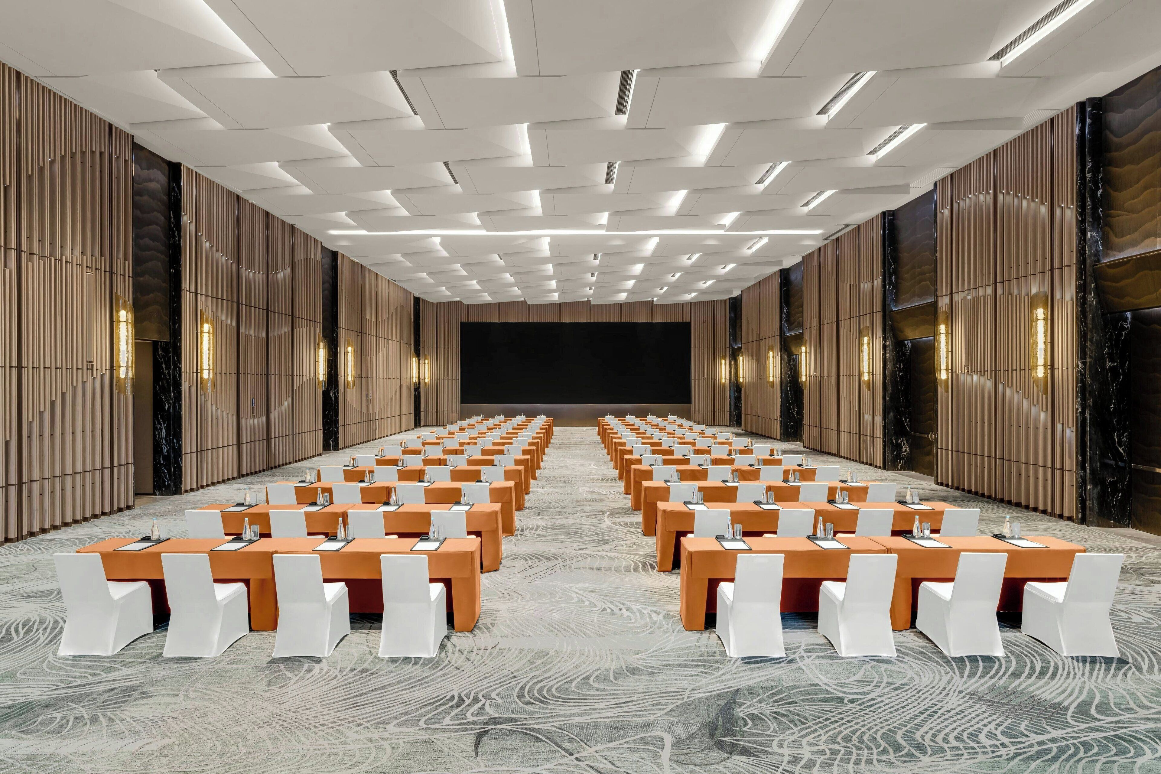 A conference room with rows of tables and chairs, a large screen at the front, and a patterned carpet.
