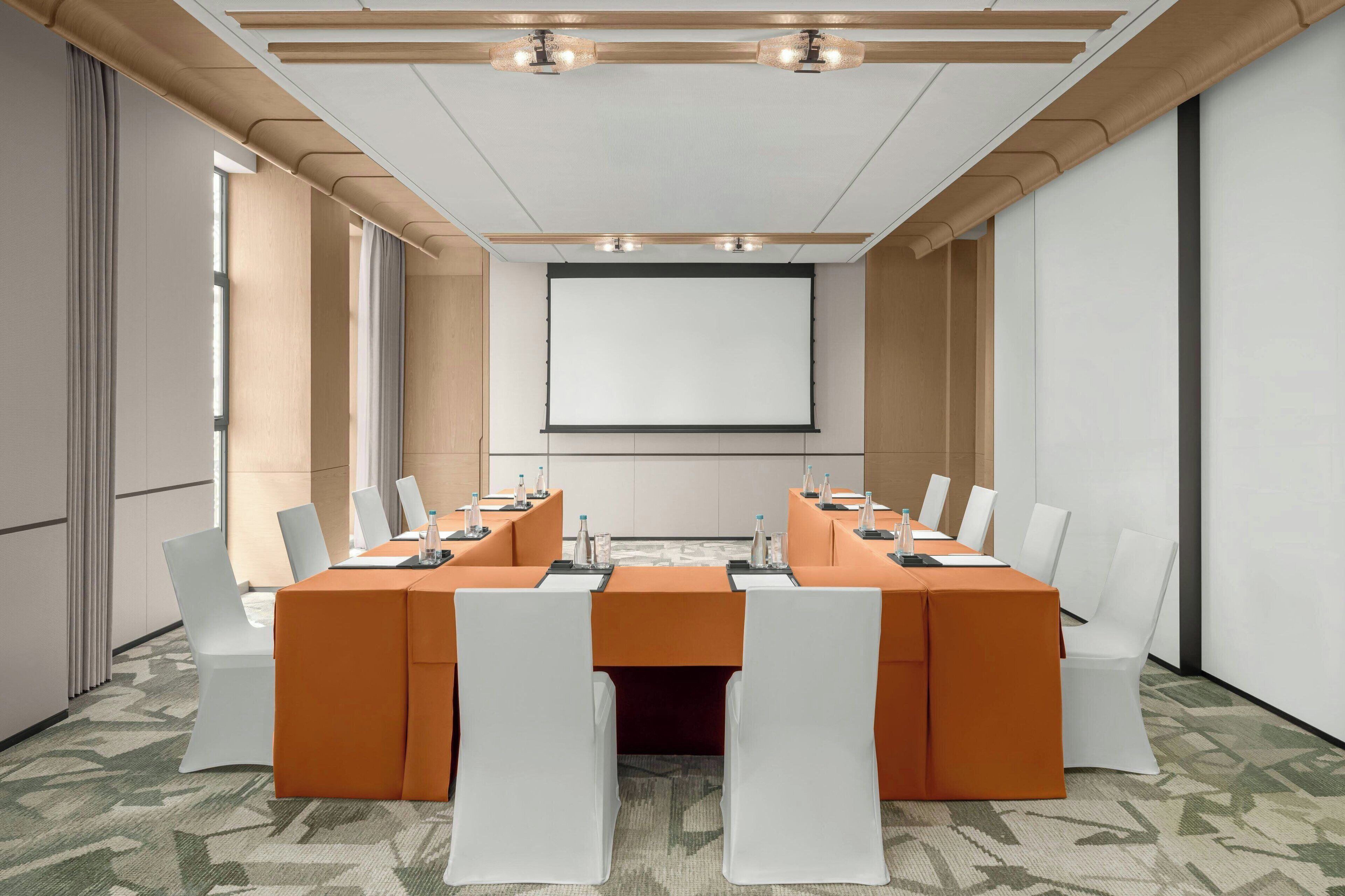 Una sala conferenze con un ampio tavolo, sedie bianche e uno schermo per proiettore.