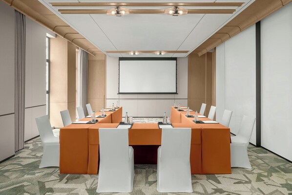 Un salón de conferencias con una mesa grande, sillas blancas y una pantalla para proyector.