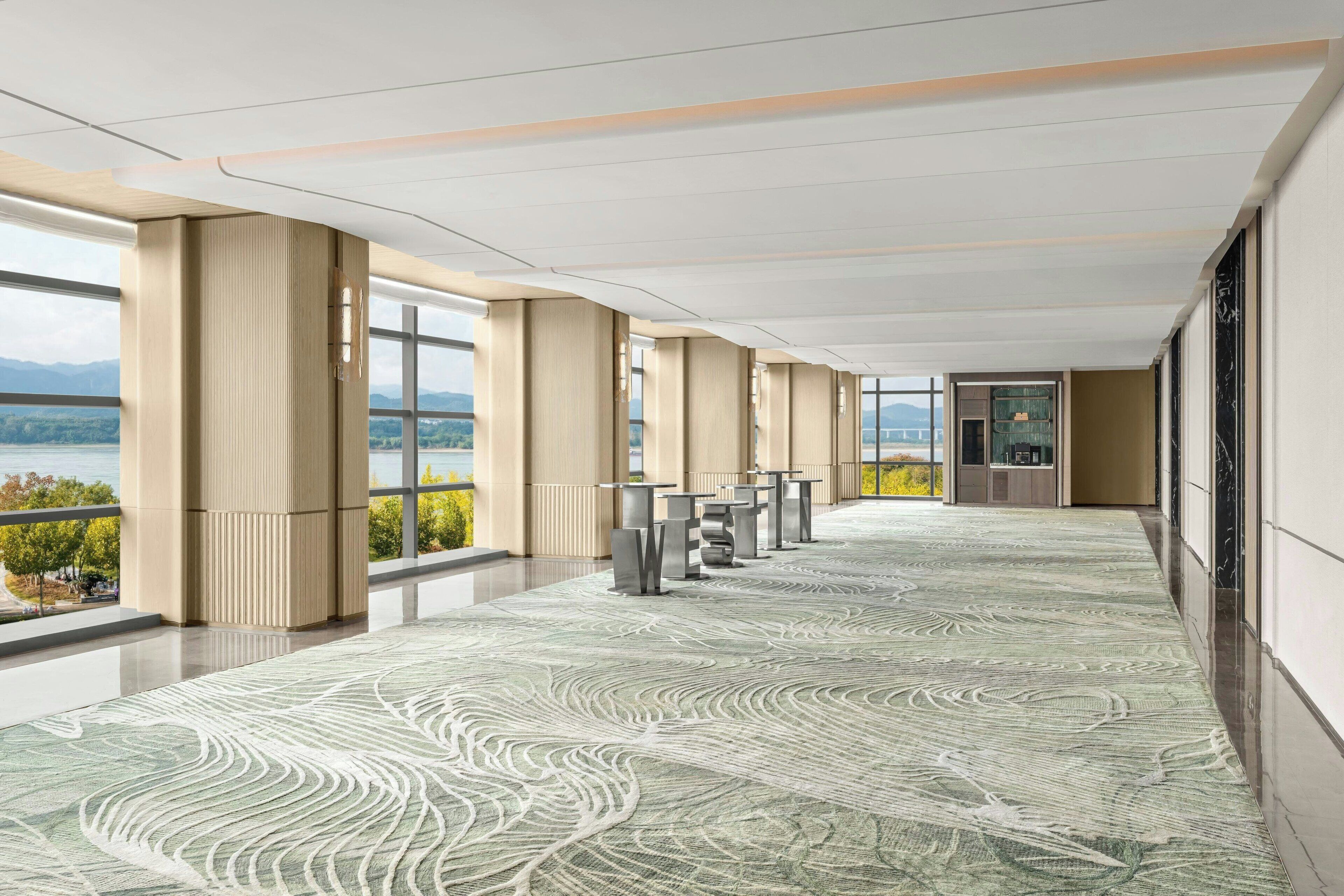 A spacious corridor with a patterned carpet, large windows offering a view of a lake and mountains, and a reception area with a glass wall.