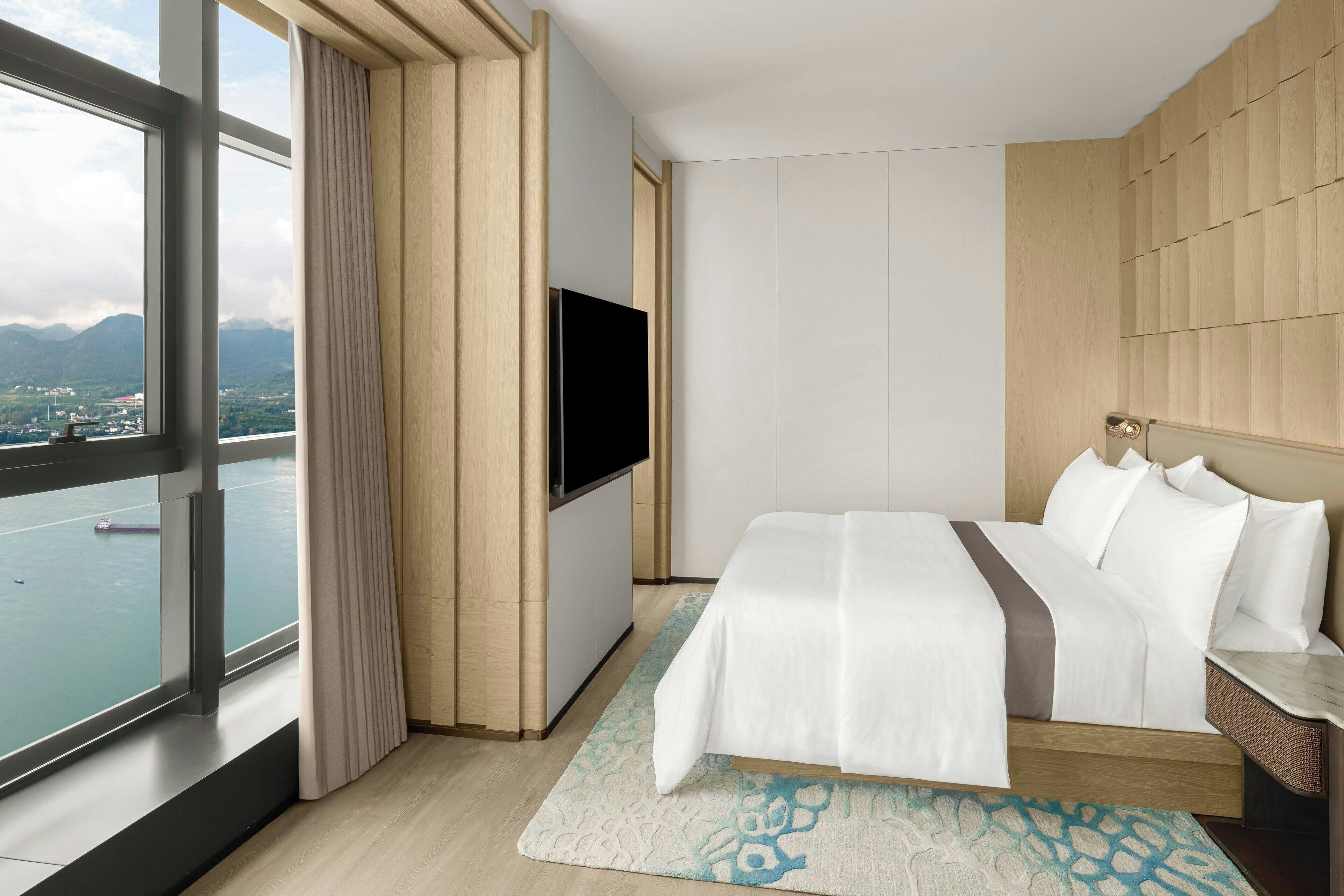 A hotel room with a large window, a bed, a TV, and a view of a body of water.