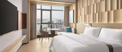A modern hotel room with a large bed, a TV, a city view, and a small table with a chair.