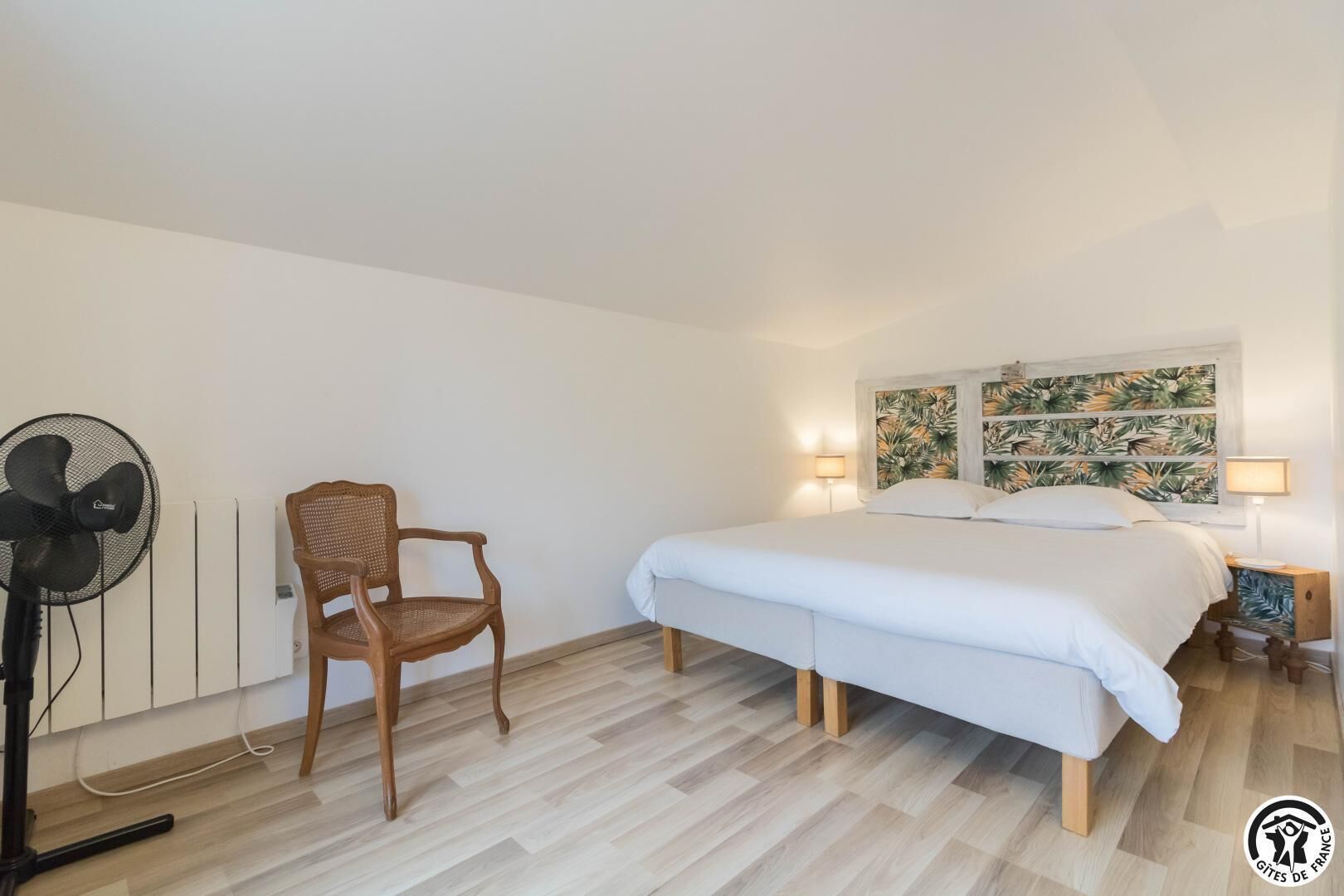 A bedroom with a bed, a chair, a standing fan, and a wall with a leafy pattern.