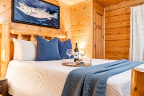 Una habitación tipo cabaña de madera con una cama, almohadas, un cuadro de un pez enmarcado, una botella de vino y copas.