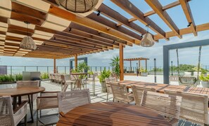 Eine Außenpatio-Terrasse mit Holztischen und Korbstühlen, eine Pergola mit hängenden Lichtern und Blick aufs Meer.