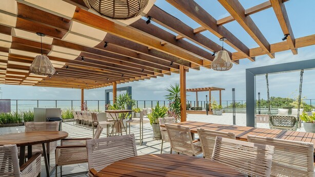 An outdoor patio area with wooden tables and wicker chairs, a pergola with hanging lights, and a view of the sea. - Beachfront Condo Studios With Pool in Bessa Beach - Gses (João Pessoa)