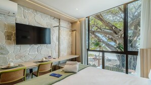 A hotel room with a stone wall, a flat-screen TV, a dining area with a table and chairs, and a large window with a view of trees and a beach. - Beachfront Condo Studios With Pool in Bessa Beach - Gses (João Pessoa)
