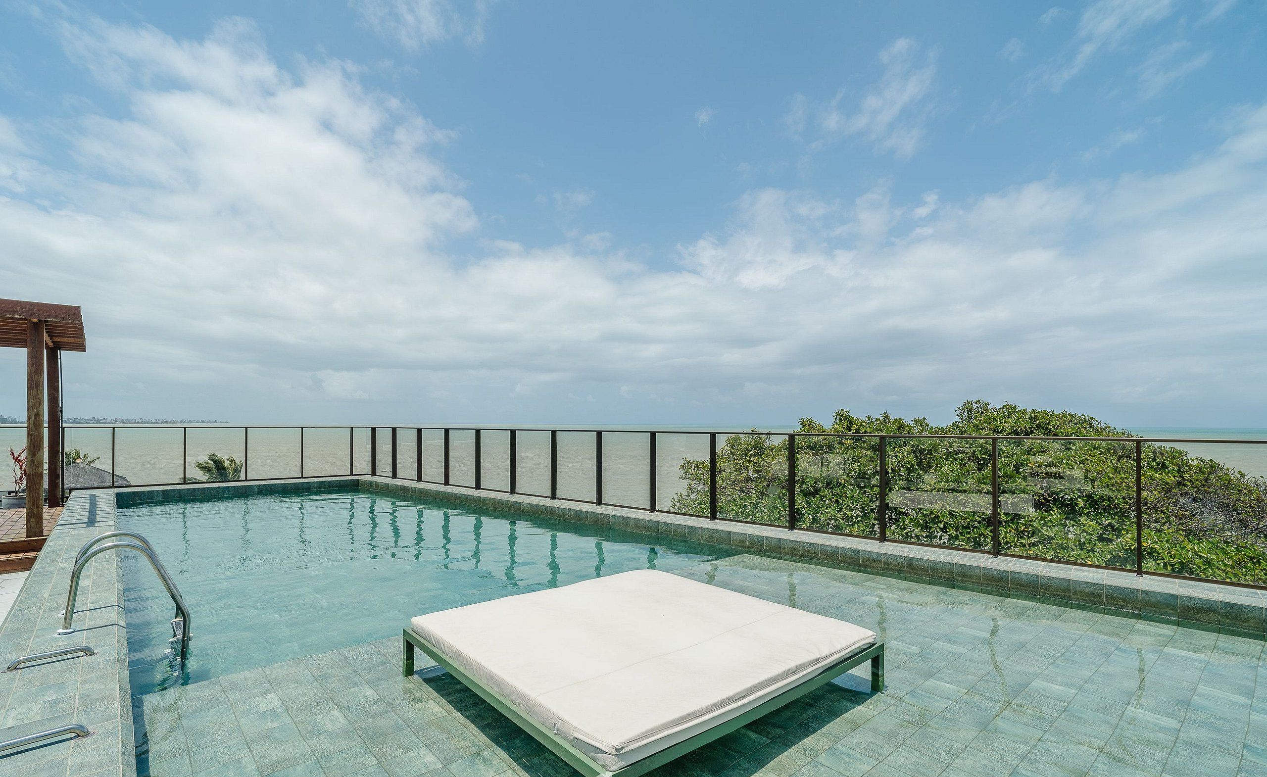 A rooftop pool with a single white lounge chair, metal railings, and a view of the sea.