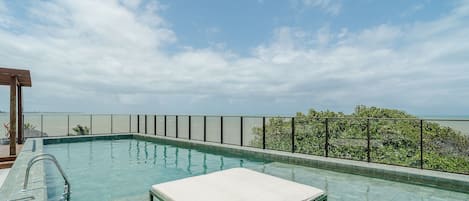 A rooftop pool with a single white lounge chair, metal railings, and a view of the sea.