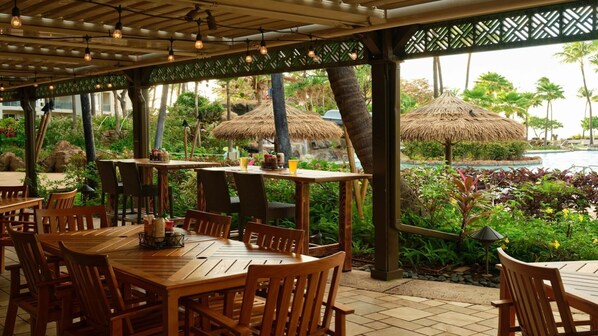 Un espace de restauration en plein air avec des tables et des chaises en bois, des parasols en chaume et une vue sur une piscine et une végétation tropicale.