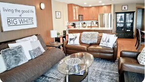 A modern living room with a brown leather sofa, a glass coffee table, and a kitchen in the background. - Raven's Reach in Happy Valley - Private Hot Tub & Shared Pool (Beaverdell)
