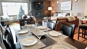 A modern living room with a dining area set for a meal. There is a fireplace, a television mounted on the wall, and a Christmas tree.