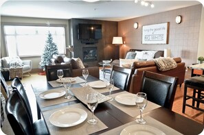 A modern living room with a dining area set for a meal. There is a fireplace, a television mounted on the wall, and a Christmas tree.