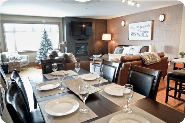A modern living room with a dining area set for a meal. There is a fireplace, a television mounted on the wall, and a Christmas tree.