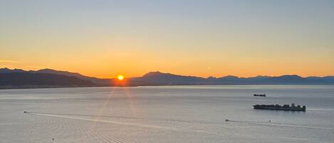A sunset over a calm body of water with ships, surrounded by mountains.