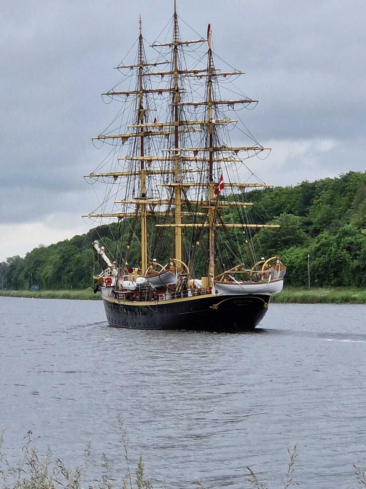 Um grande navio a vela com vários mastros e cabos, navegando em um corpo d'água com uma costa arborizada ao fundo.