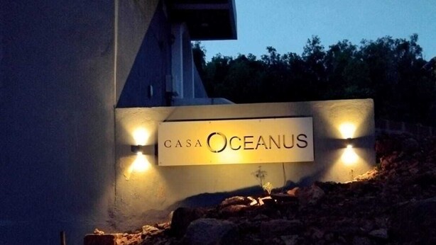A sign for 'Casa Oceanus' with two wall-mounted lights, a staircase leading up to the building, and a rocky terrain. - Casa Oceanus - 3 Bedroom - Sea Facing Pool Villa - Ganpatipule - Ratnagiri (Ganpatipule - Ratnagiri)
