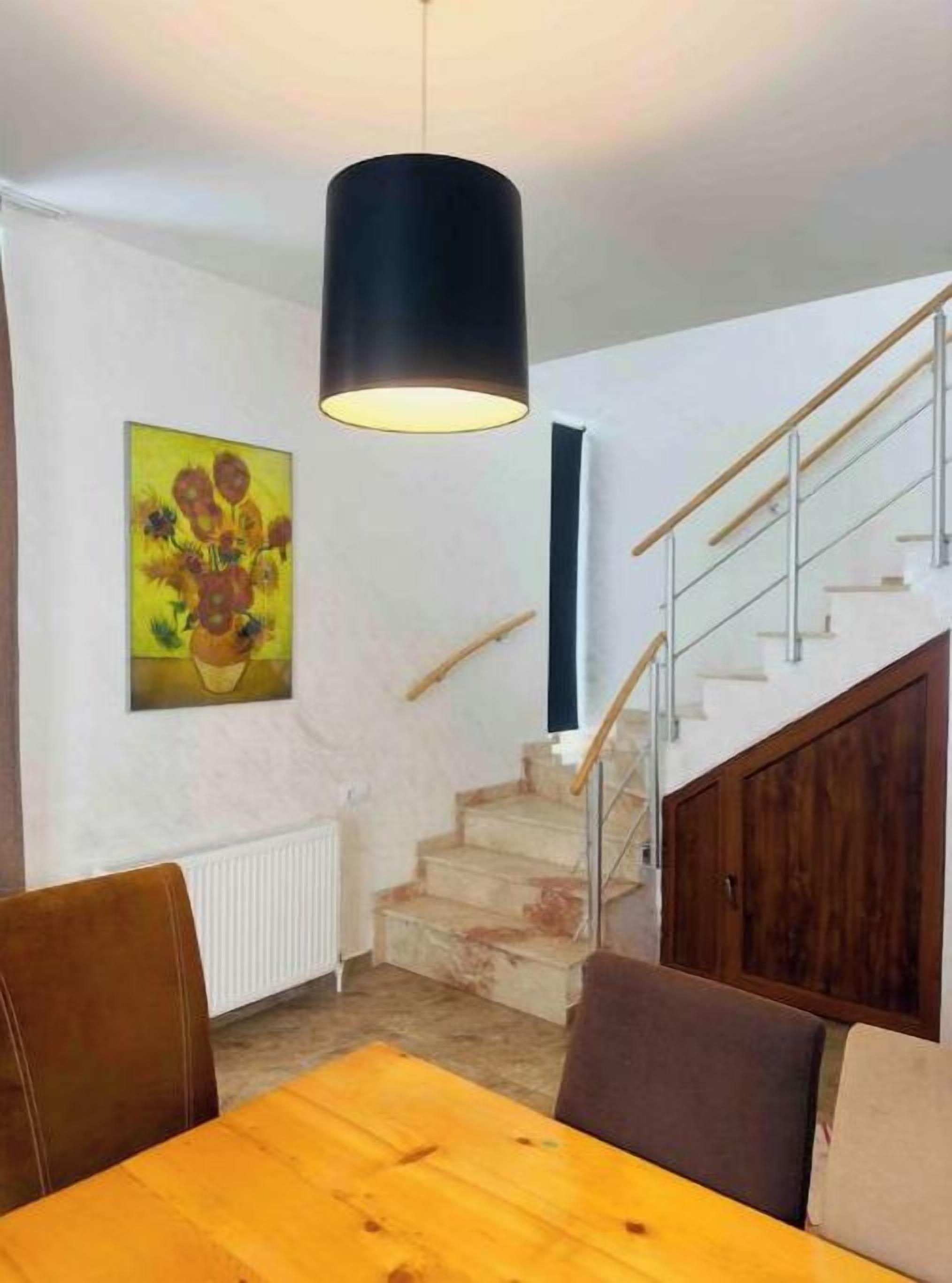 A dining area with a wooden table and chairs, a black pendant light, a staircase with wooden railings, and a painting on the wall.