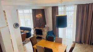 Un salon avec une cheminée, une table à manger avec des chaises, un canapé et une grande fenêtre avec des rideaux.
