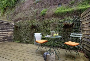 Eine Holzterrasse mit Tisch und zwei Stühlen, umgeben von einer moosbewachsenen Mauer.