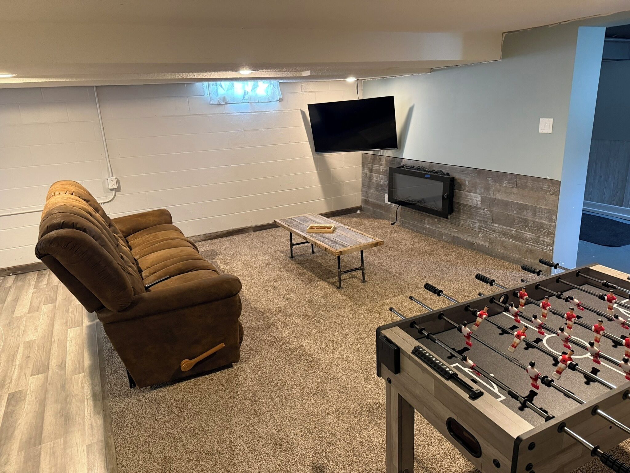 Een gezellige woonkamer met een bruine bank, een tafelvoetbaltafel, een bijzettafeltje en een flatscreen televisie aan de muur.