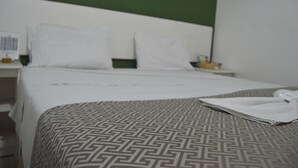 A hotel room with a bed, white pillows, a patterned bedspread, and a nightstand with a lamp and a bottle. - Hotel Cuca Fresca (Cotia)