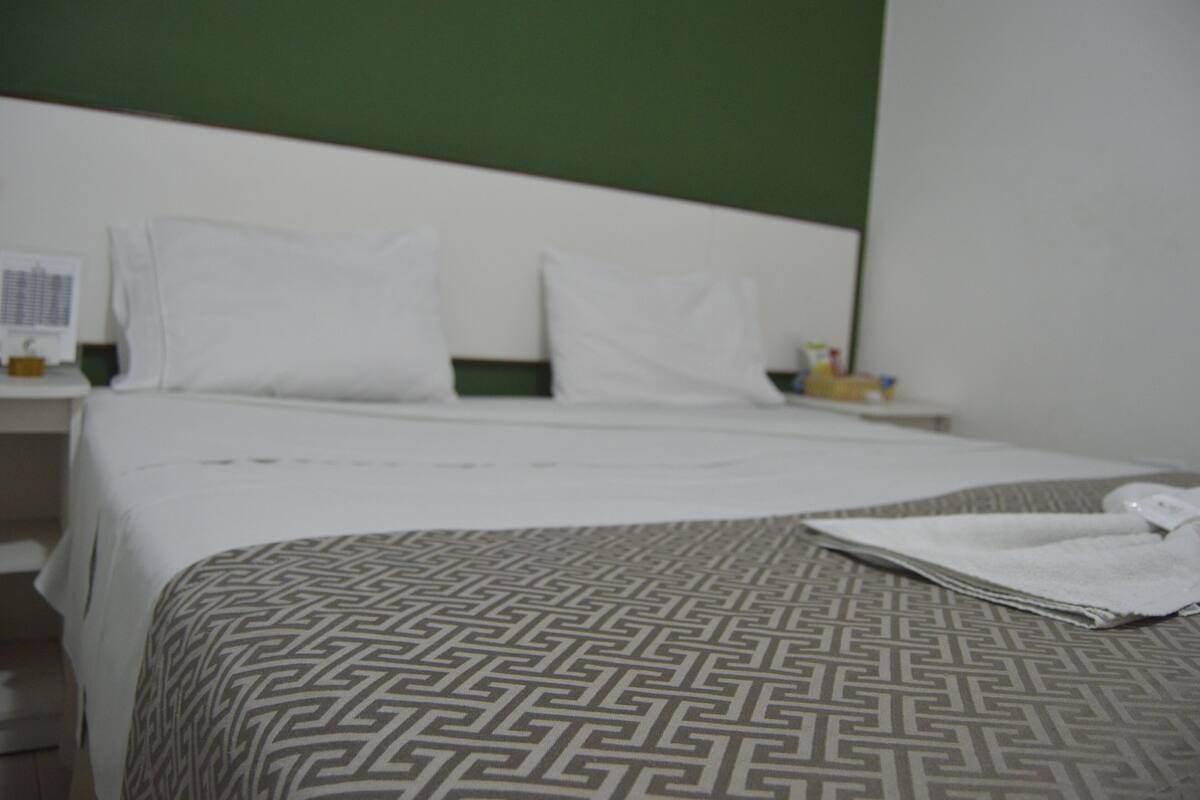 A hotel room with a bed, white pillows, a patterned bedspread, and a nightstand with a lamp and a bottle.