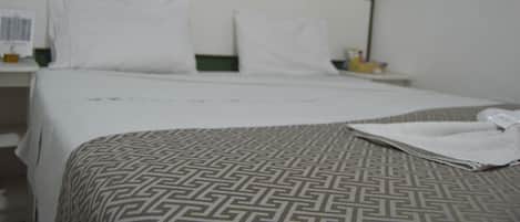 Habitación de hotel con cama, almohadas blancas, colcha estampada y mesita de noche con lámpara y botella.