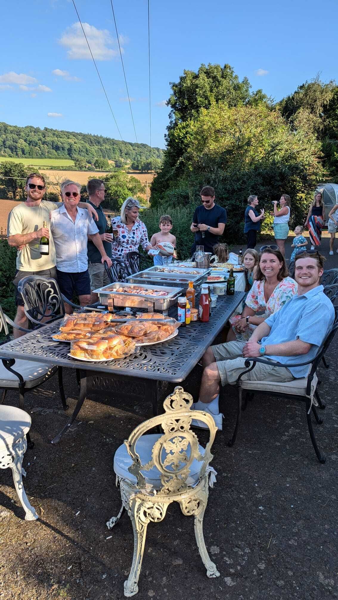 Un grupo de personas reunidas alrededor de una mesa con comida y bebidas, al aire libre con una vista panorámica de fondo.