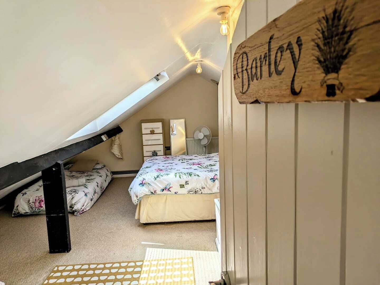 Ein gemütliches Schlafzimmer im Dachgeschoss mit schrägen Wänden, einer Holzschrifttafel, einem Bett mit floralem Bettzeug, einem Schminktisch und einem Ventilator.