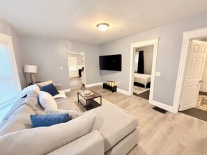A modern living room with a sofa, a coffee table, and a TV. There is a hallway leading to a bedroom with a bed. - Luxe charming  2-bedroom apartment in Rochester (Rochester)