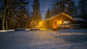 Une cabane dont la véranda et les marches menant à l’entrée sont recouvertes de neige, entourée d’arbres et d’un paysage enneigé.