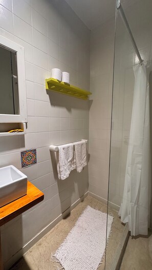 Une salle de bain avec des carreaux blancs, une douche avec une porte en verre, un lavabo blanc, une étagère en bois avec des serviettes et une petite mosaïque décorative sur le mur.