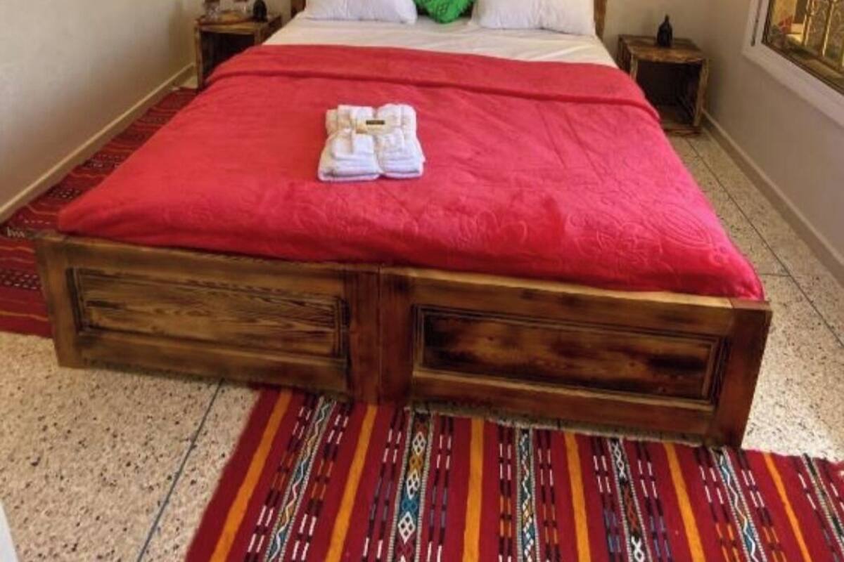 A large bed with a red bedspread, a wooden headboard, and a green pillow. There is a painting on the wall above the bed and a patterned rug on the floor.