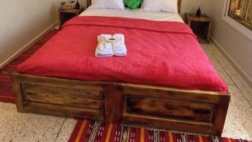 A large bed with a red bedspread, a wooden headboard, and a green pillow. There is a painting on the wall above the bed and a patterned rug on the floor.