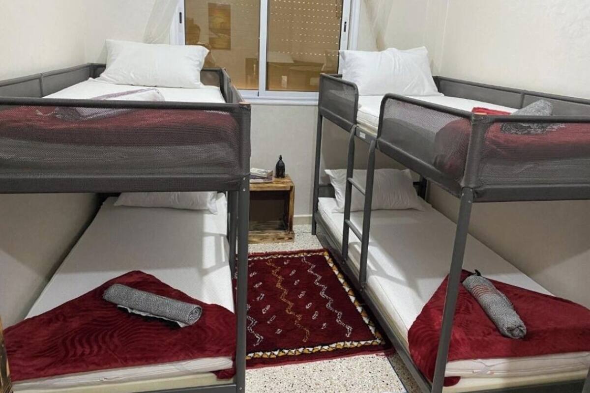 A room with three bunk beds, each with a red blanket and a rolled-up towel. There is a small wooden nightstand with a lamp between the beds.